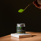 Jar of matcha green tea powder with a spoonful being held above it on a dark background