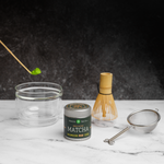 Matcha tea gift set with a tin of Mizuba Okumidori single cultivar matcha. A bamboo chashaku spoon  with matcha hovers over a glass chawan tea bowl that's next to a bamboo chasen whisk and metal sieve on a marble surface.