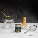 Matcha tea gift set with a tin of Mizuba Okumidori single cultivar matcha. A bamboo chashaku spoon  with matcha hovers over a glass chawan tea bowl that's next to a bamboo chasen whisk and metal sieve on a marble surface.