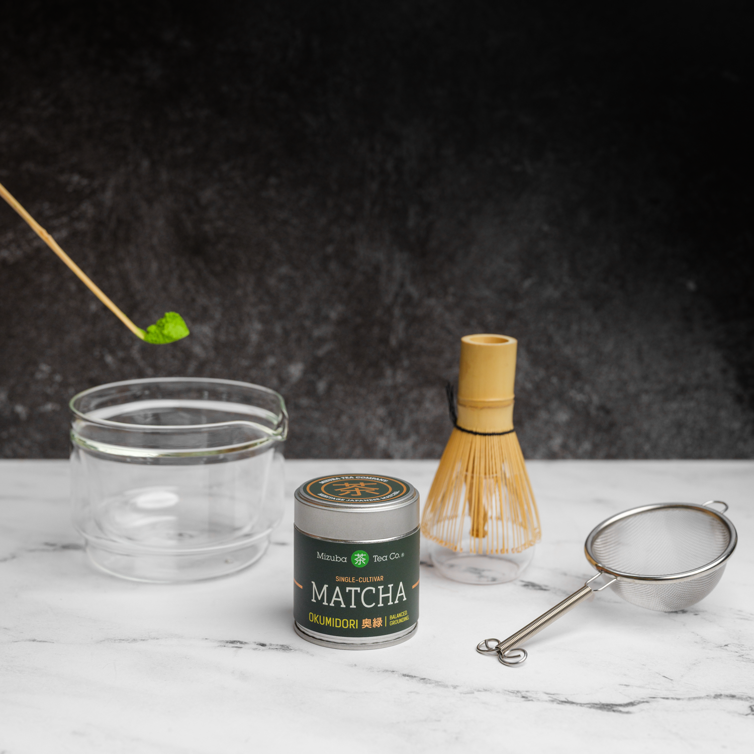 Matcha tea gift set with a tin of Mizuba Okumidori single cultivar matcha. A bamboo chashaku spoon  with matcha hovers over a glass chawan tea bowl that's next to a bamboo chasen whisk and metal sieve on a marble surface.