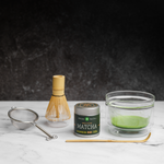 Matcha green tea set complete with traditional tools. A glass chawan bowl with frothy matcha green tea inside sits next to a whisk,  a bamboo chashaku scoop, a metal sieve, and a Mizuba Tea Okumidori matcha tin on a dark background.