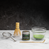 Matcha green tea set complete with traditional tools. A glass chawan bowl with frothy matcha green tea inside sits next to a whisk,  a bamboo chashaku scoop, a metal sieve, and a Mizuba Tea Okumidori matcha tin on a dark background.