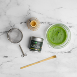 Hario glass Mizuba Matcha green tea set with a tin of single-cultivar Okumidori, whisk, metal sieve, and bamboo spoon on a marble surface.