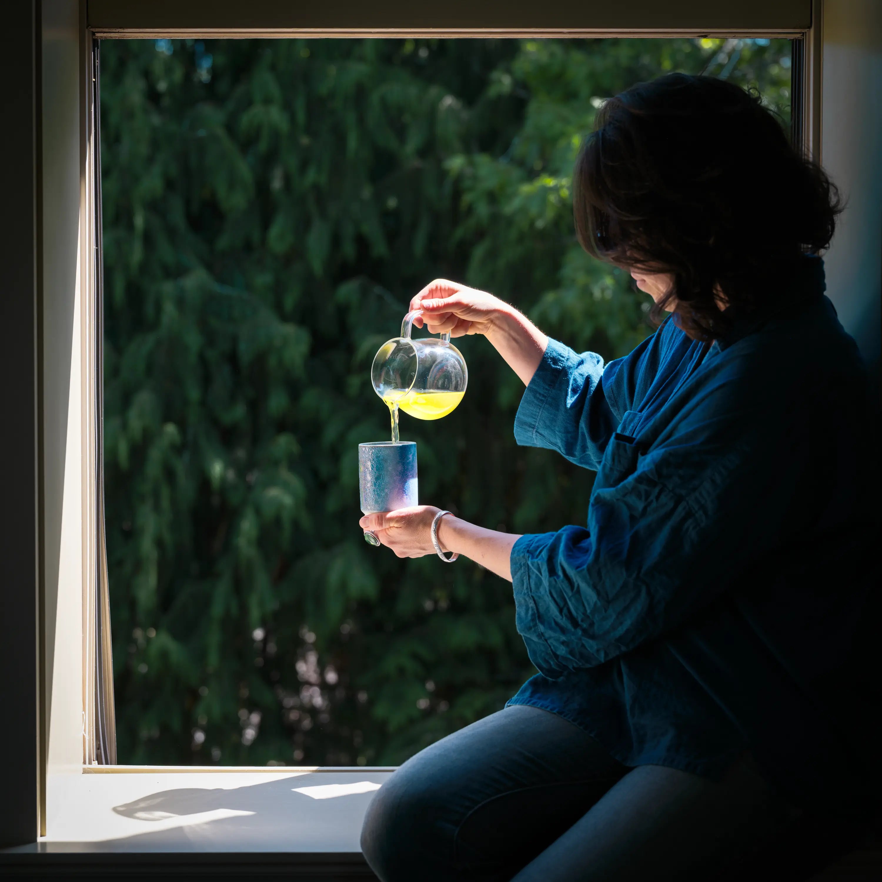 A woman pours freshly brewed shincha Japanese green tea into a blue ceramic cup. 