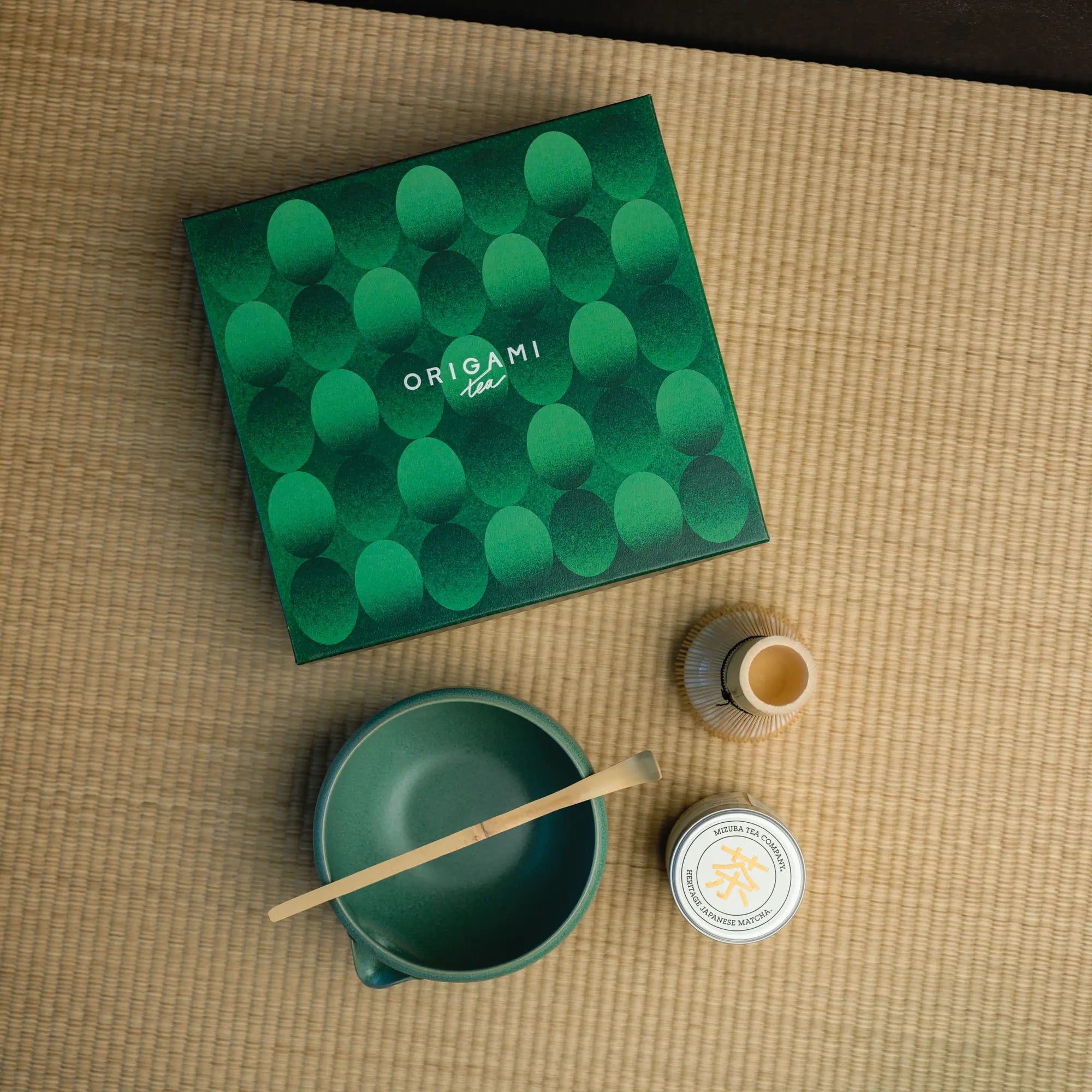 Mizuba Matcha Tin A chasen, chawan, and matcha bowl in front of a green box labeled "Origami Tea"