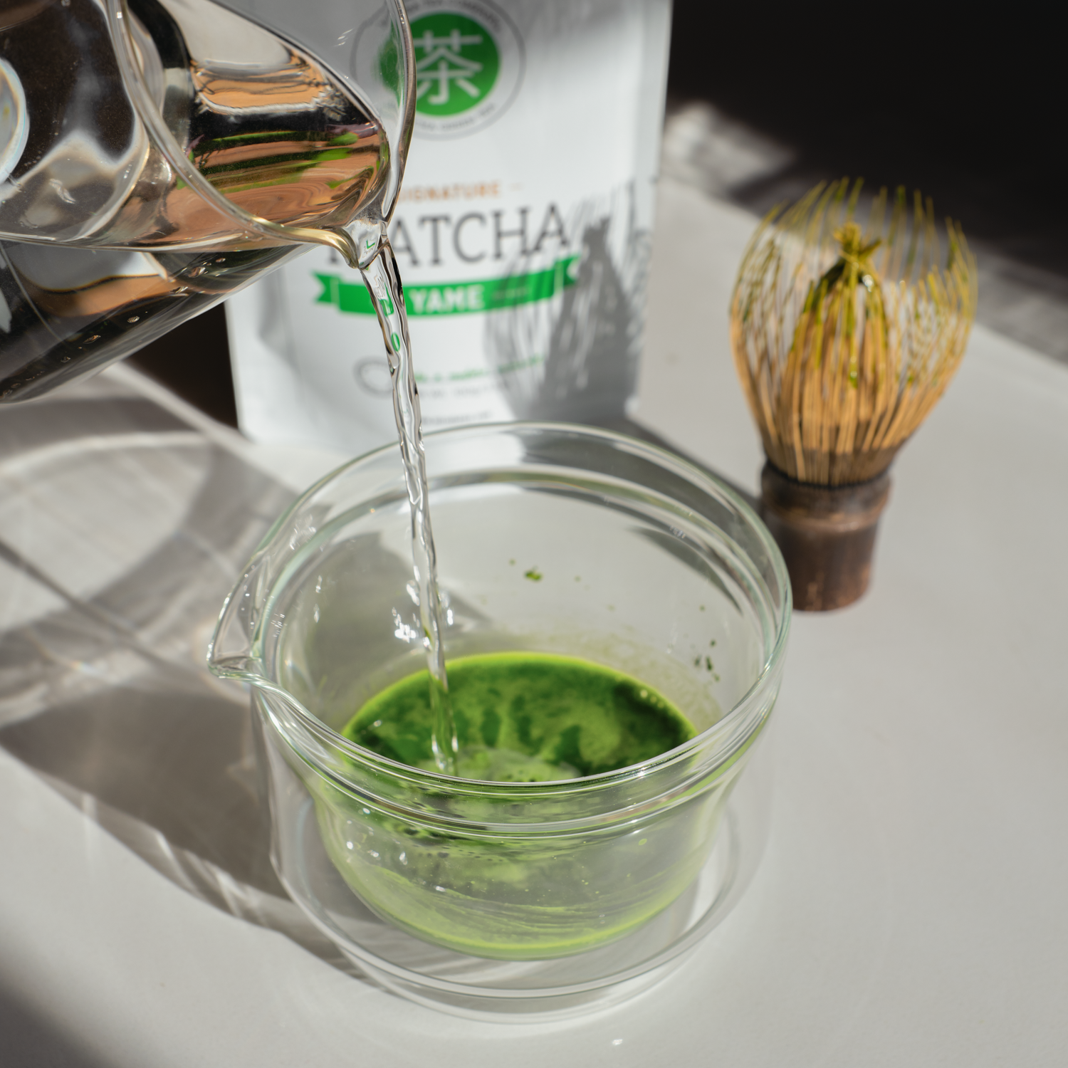 Water intended to make matcha is poured into a glass chawan (tea bowl) filled with Yame First Flush matcha green tea with its packaging in the background. 