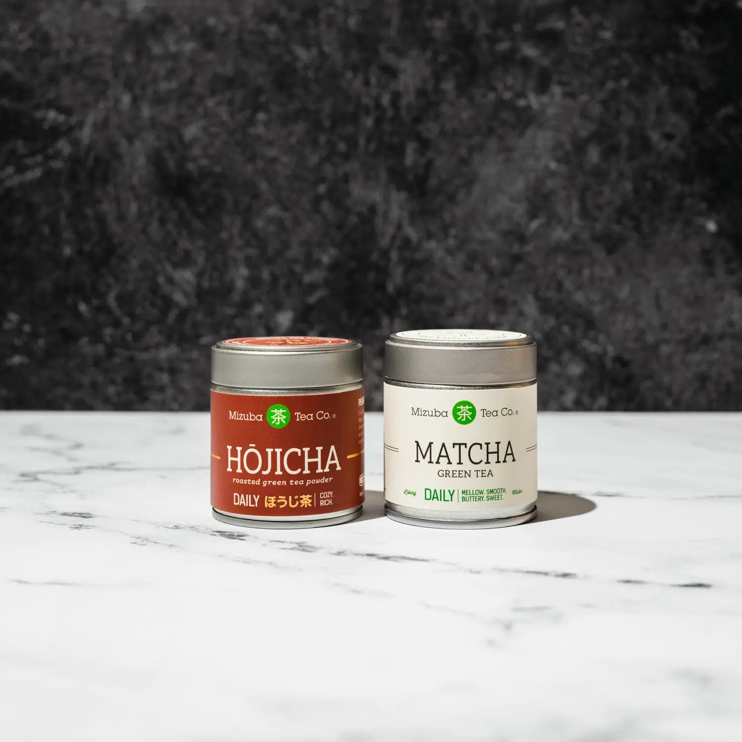 Two Mizuba tea canisters, one labeled 'Hojicha' and the other 'Matcha', on a marble surface with a dark textured background.